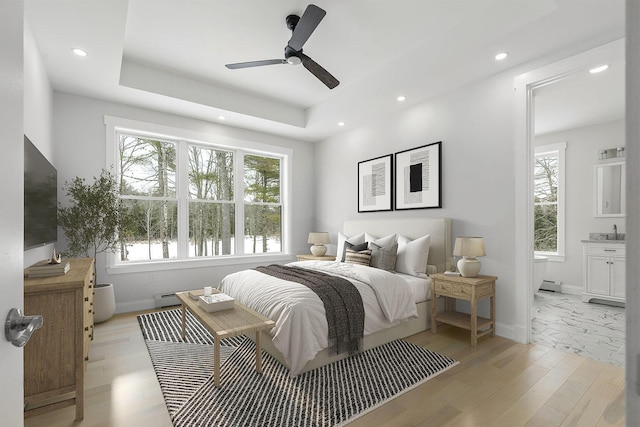 bedroom with ensuite bath, ceiling fan, baseboard heating, a tray ceiling, and light wood-type flooring