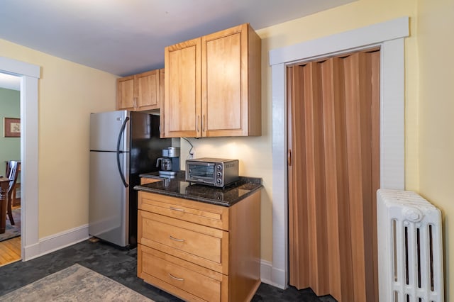 kitchen with light brown cabinetry, radiator, stainless steel fridge, and dark stone counters