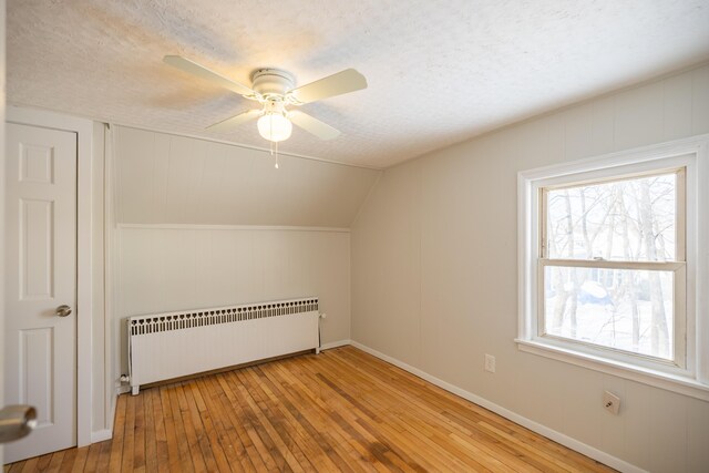 additional living space with lofted ceiling, ceiling fan, radiator heating unit, light wood-style flooring, and a textured ceiling