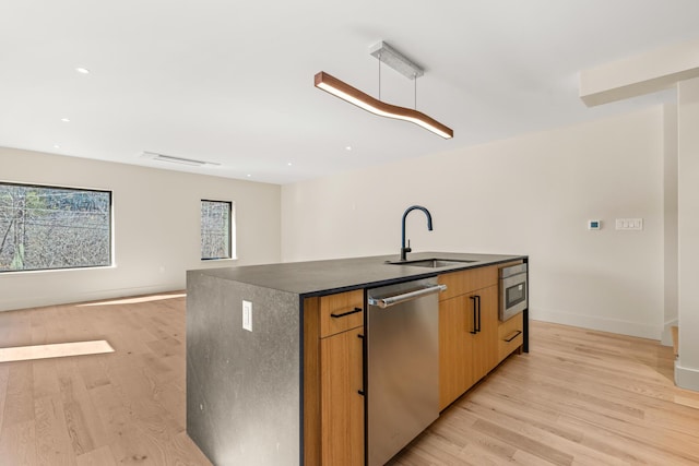 kitchen featuring light wood-style flooring, a kitchen island with sink, a sink, dark countertops, and appliances with stainless steel finishes