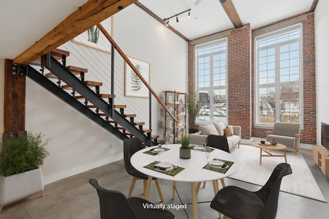 dining area featuring rail lighting, beam ceiling, a towering ceiling, brick wall, and concrete floors