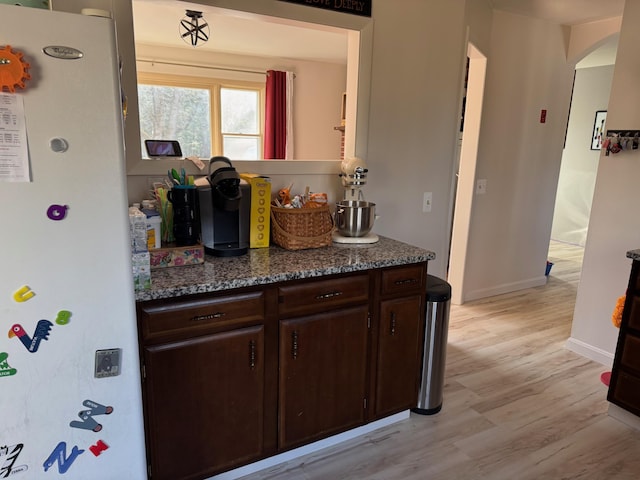 kitchen featuring freestanding refrigerator, arched walkways, light wood-style floors, dark brown cabinetry, and baseboards