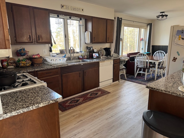 kitchen with light wood finished floors, dark brown cabinetry, dark stone counters, white appliances, and a sink