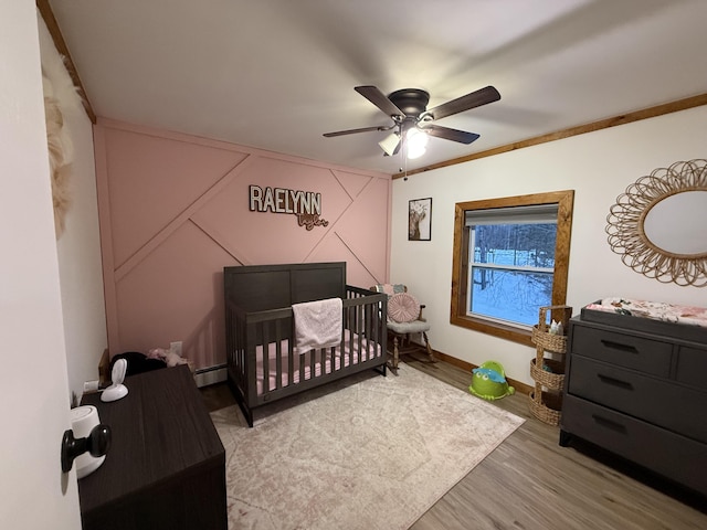 bedroom featuring a nursery area, hardwood / wood-style flooring, ceiling fan, baseboard heating, and crown molding