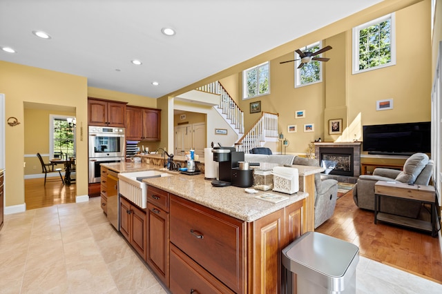 kitchen featuring sink, light stone counters, a center island with sink, ceiling fan, and stainless steel double oven