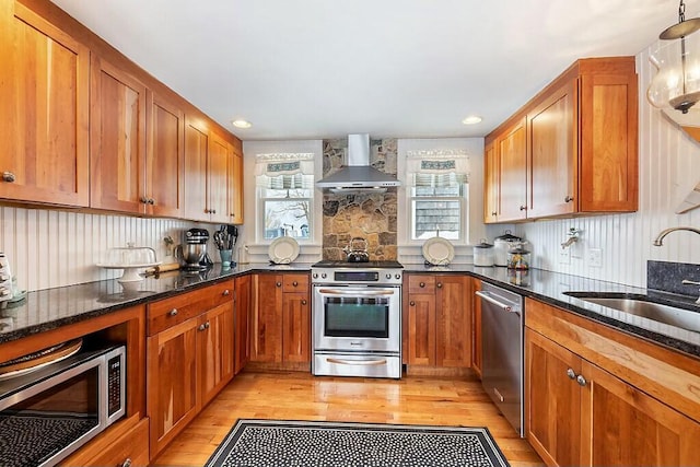 kitchen with brown cabinets, stainless steel appliances, a sink, dark stone countertops, and wall chimney exhaust hood