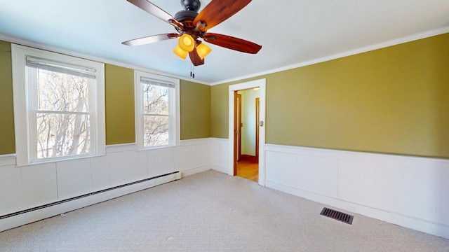 carpeted empty room featuring visible vents, a wainscoted wall, crown molding, baseboard heating, and ceiling fan