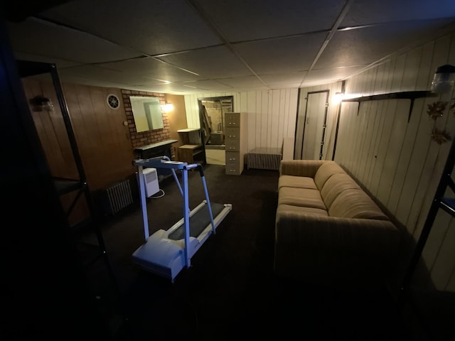 basement featuring a paneled ceiling, radiator heating unit, a brick fireplace, and wood walls