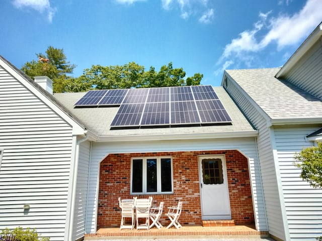 doorway to property featuring roof with shingles, roof mounted solar panels, a chimney, and brick siding