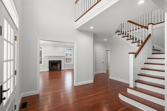 foyer entrance with dark wood-style flooring, a fireplace with flush hearth, visible vents, a towering ceiling, and baseboards