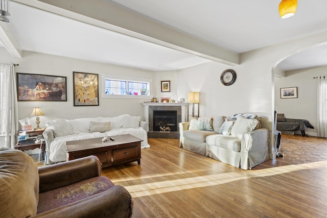 living area featuring arched walkways, beamed ceiling, a tiled fireplace, and light wood-style floors