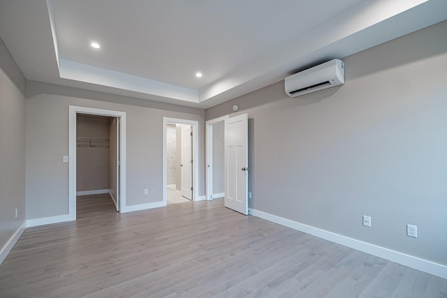 unfurnished bedroom with a raised ceiling, a walk in closet, a wall mounted air conditioner, and light wood-type flooring