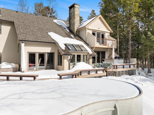 snow covered property with a balcony, a chimney, a deck, and roof with shingles