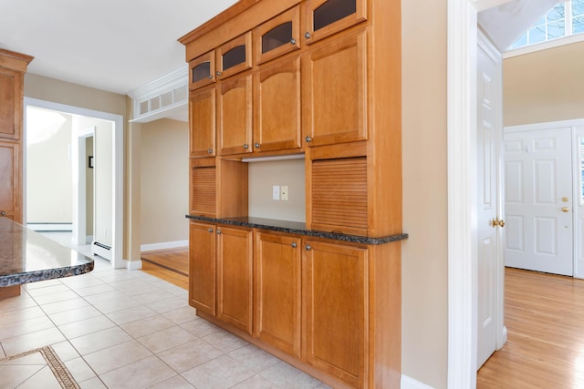 kitchen featuring brown cabinets, a baseboard radiator, glass insert cabinets, light tile patterned flooring, and dark stone countertops