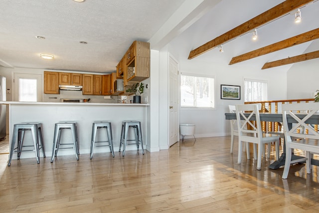 kitchen featuring beam ceiling, brown cabinets, light wood-style flooring, under cabinet range hood, and a kitchen breakfast bar
