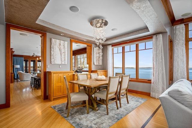 dining area with a chandelier, light wood-style flooring, a water view, baseboards, and a tray ceiling