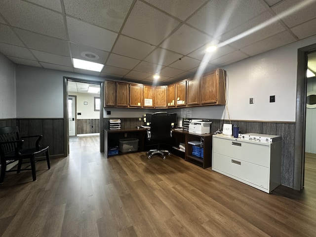 home office with dark wood finished floors, a drop ceiling, built in study area, wainscoting, and wood walls