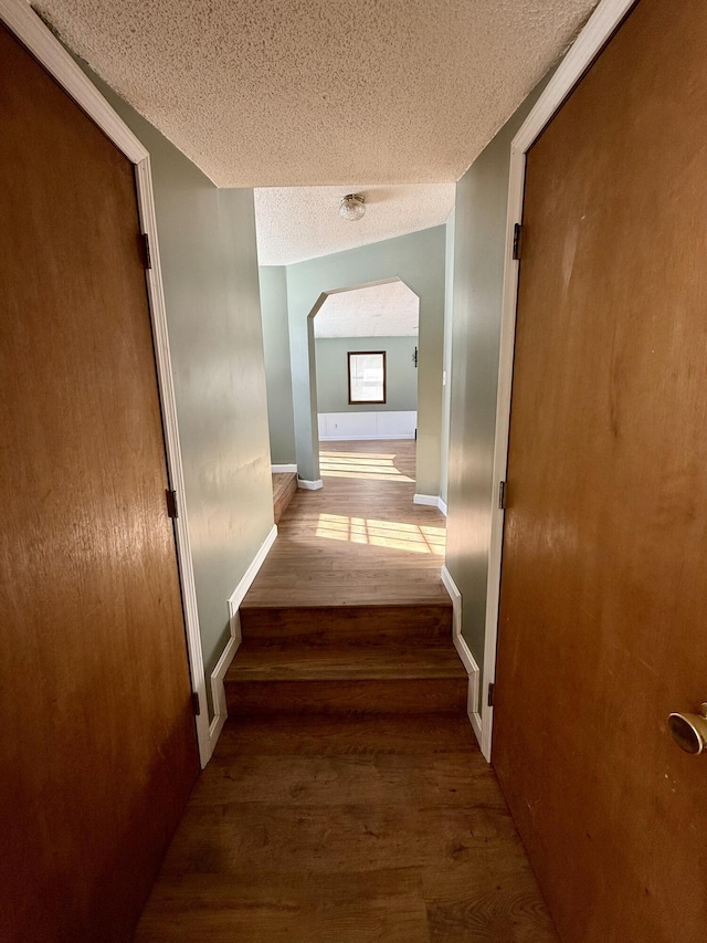 hallway featuring a textured ceiling, arched walkways, wood finished floors, and baseboards