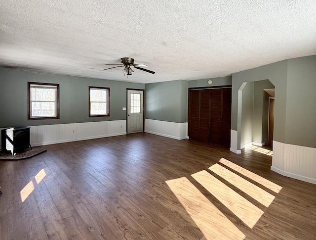 unfurnished living room featuring a wood stove, a wainscoted wall, dark wood-style floors, and a wealth of natural light