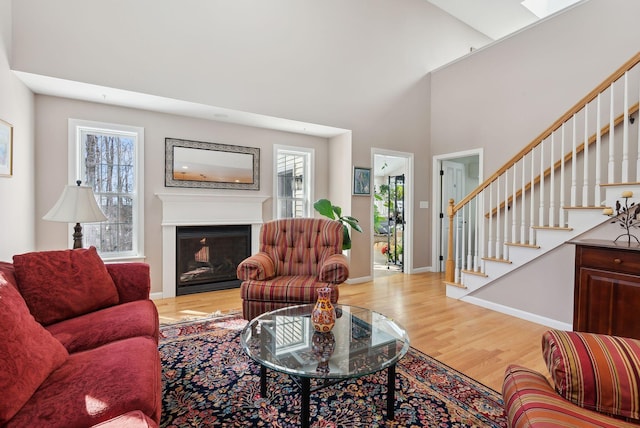 living area with a towering ceiling, a glass covered fireplace, light wood-type flooring, baseboards, and stairs