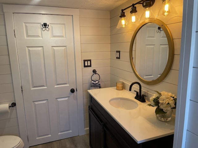 half bathroom with a textured ceiling, toilet, wooden walls, wood finished floors, and vanity