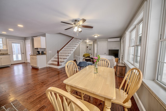 dining room featuring a wall unit AC, dark wood finished floors, stairway, a ceiling fan, and baseboards