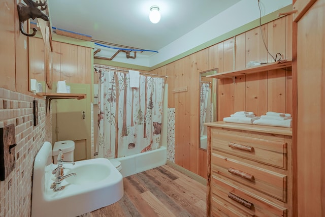 bathroom with toilet, wooden walls, a sink, wood finished floors, and shower / bathtub combination with curtain