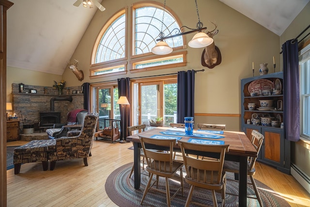 dining space with a ceiling fan, a baseboard radiator, a wood stove, light wood-type flooring, and high vaulted ceiling