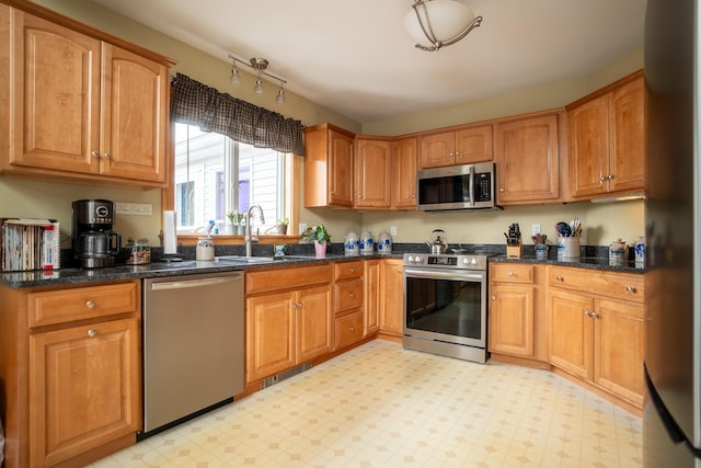 kitchen with light floors, appliances with stainless steel finishes, brown cabinetry, a sink, and dark stone counters