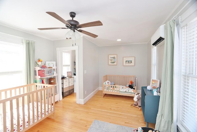 bedroom with ceiling fan, a wall unit AC, baseboards, light wood-style floors, and a nursery area