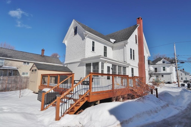 snow covered back of property featuring an outbuilding, fence, a sunroom, a wooden deck, and a chimney