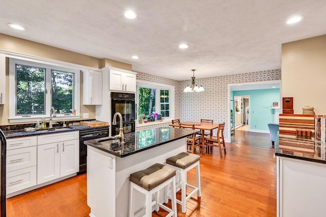 kitchen with a kitchen island with sink, a sink, white cabinets, hanging light fixtures, and wallpapered walls