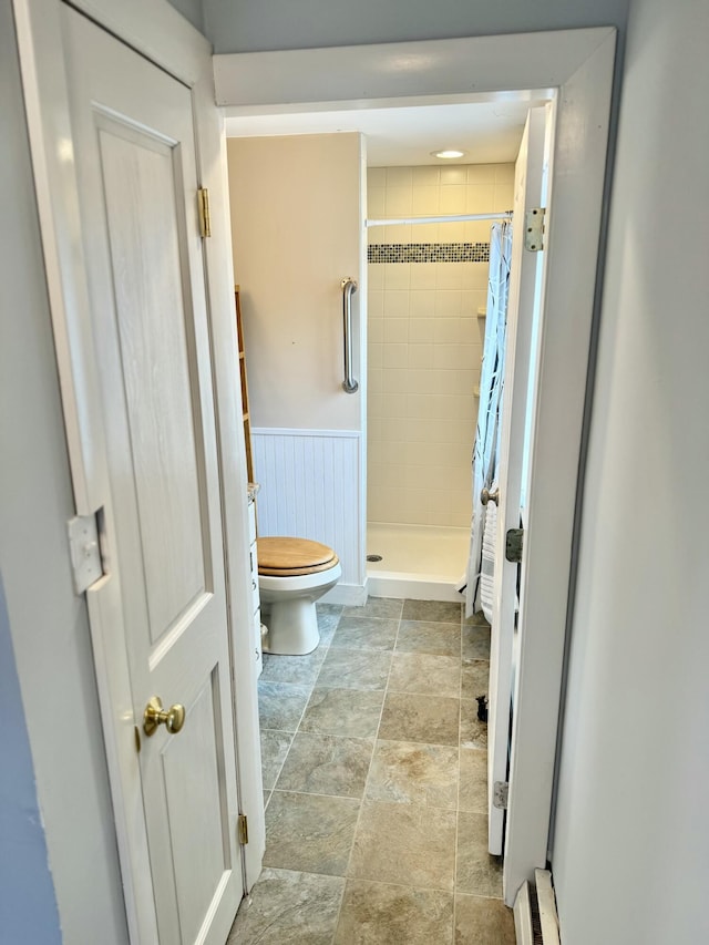 full bath with toilet, a stall shower, baseboard heating, wainscoting, and stone finish floor