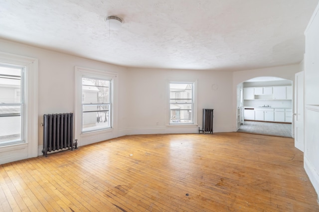 unfurnished living room with arched walkways, a textured ceiling, baseboards, light wood-type flooring, and radiator