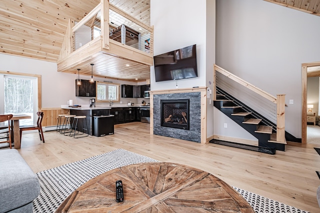 living area featuring a glass covered fireplace, wooden ceiling, light wood-style flooring, stairs, and high vaulted ceiling