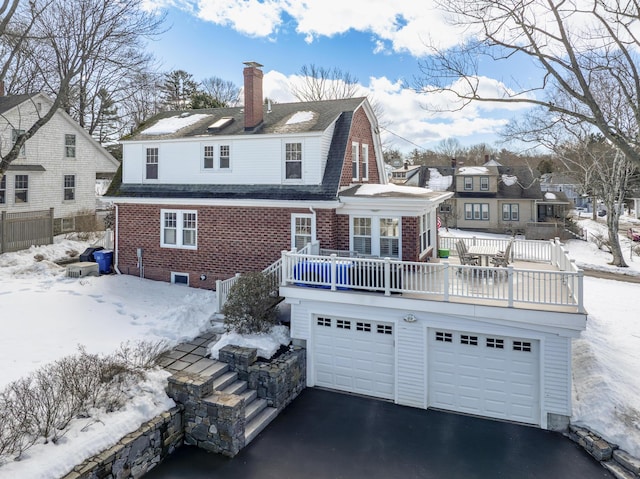 snow covered house with driveway, a gambrel roof, a chimney, a deck, and brick siding