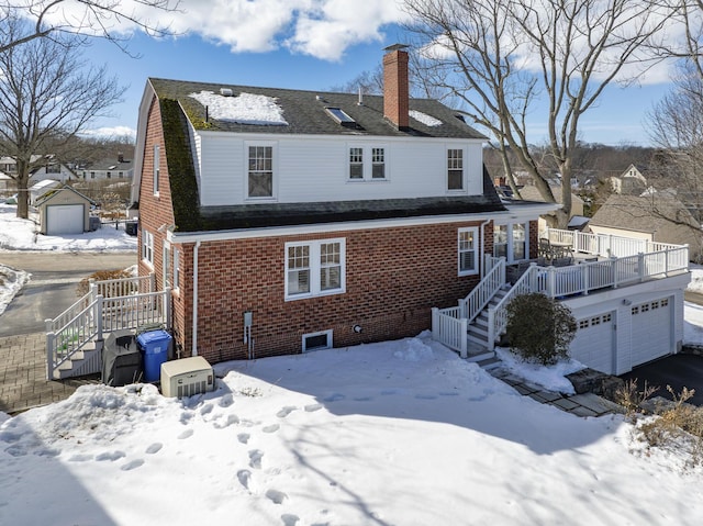 snow covered back of property featuring brick siding, roof with shingles, a chimney, a deck, and a garage