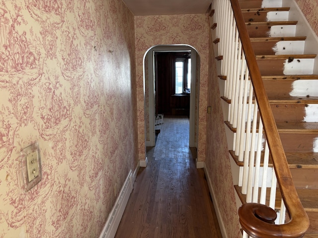 hallway featuring arched walkways, dark wood-type flooring, baseboards, stairs, and wallpapered walls