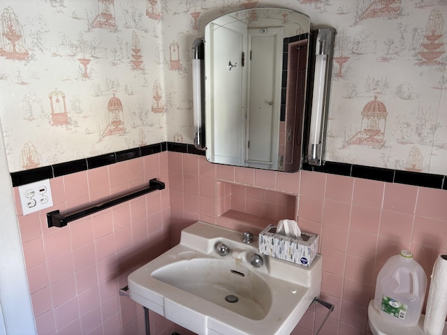 bathroom featuring wallpapered walls, tile walls, a sink, and wainscoting