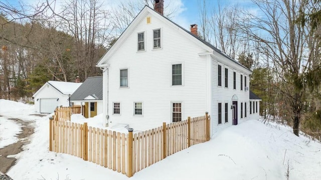 snow covered back of property featuring a detached garage, a chimney, and fence