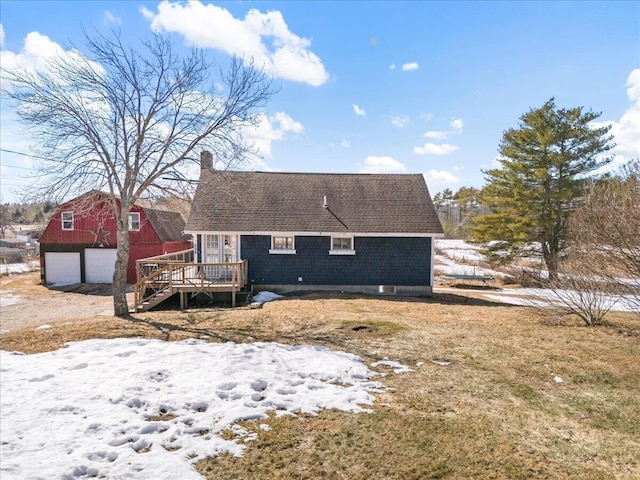 snow covered back of property with a wooden deck, roof with shingles, a chimney, an outdoor structure, and a garage