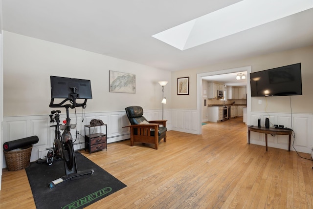 workout area featuring a skylight, a baseboard heating unit, light wood finished floors, and a wainscoted wall