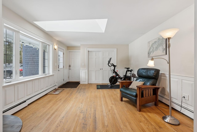 living area with a skylight, a baseboard radiator, light wood-style floors, and a wainscoted wall