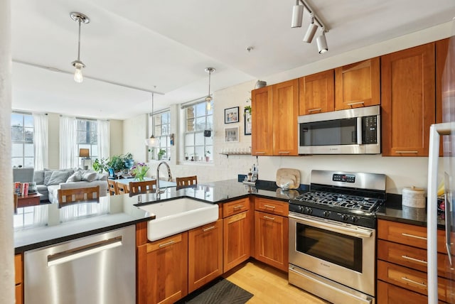 kitchen with stainless steel appliances, a sink, brown cabinets, dark countertops, and pendant lighting