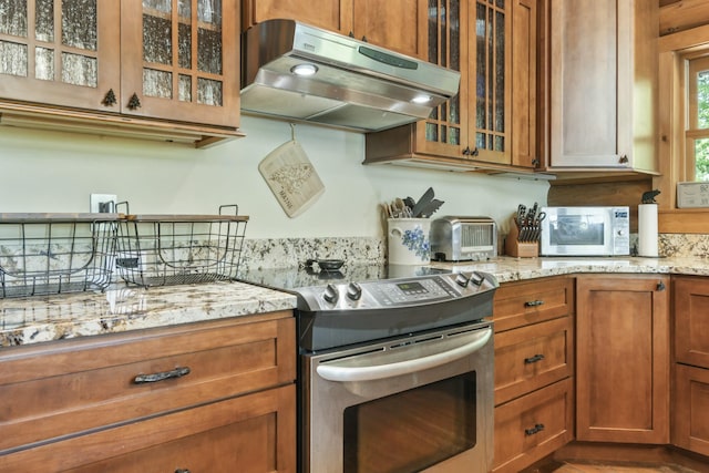 kitchen featuring brown cabinets, glass insert cabinets, stainless steel range with electric cooktop, light stone countertops, and under cabinet range hood