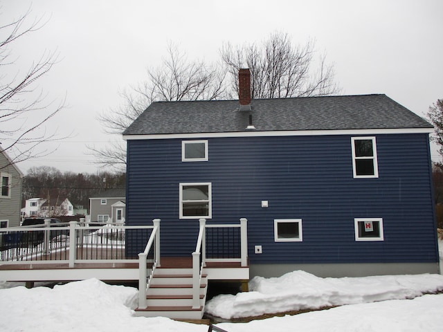 snow covered house with roof with shingles, a chimney, and a wooden deck