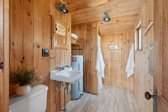 half bathroom with a sink, toilet, wood walls, water heater, and wood finished floors