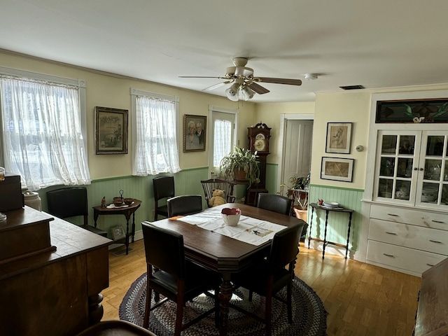 dining room with light wood finished floors, a wainscoted wall, visible vents, and a ceiling fan