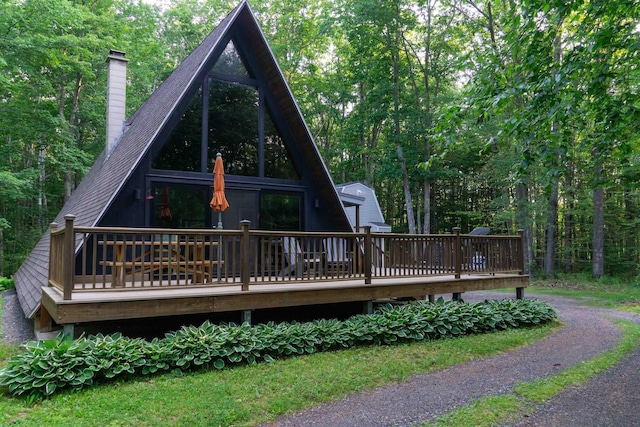 rear view of house featuring a deck, driveway, roof with shingles, a chimney, and a wooded view