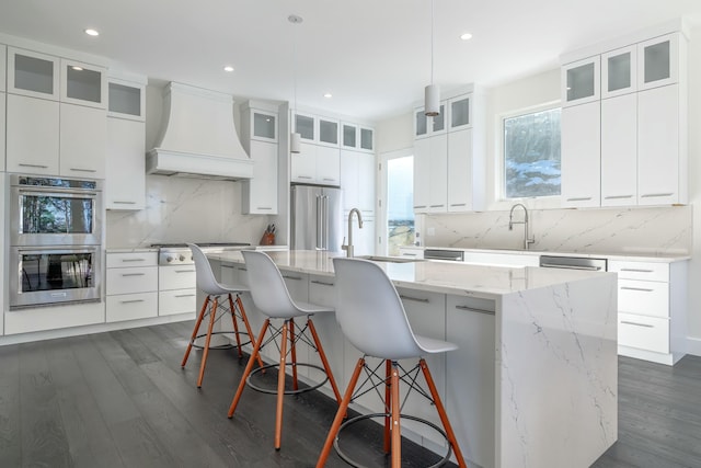 kitchen with dark wood-style flooring, stainless steel appliances, a kitchen island with sink, white cabinetry, and premium range hood